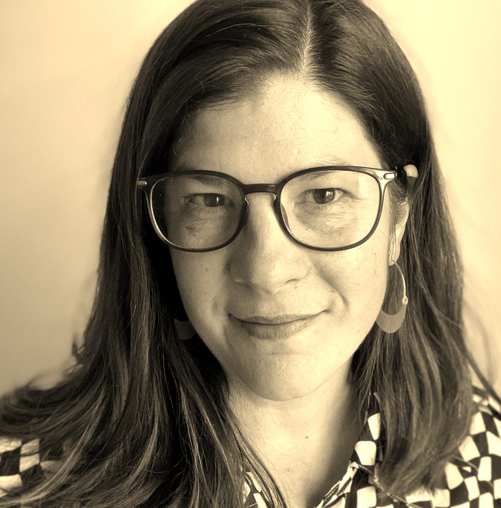 A close-up photo sepia toned image of a white, brunette woman in glasses. She is smiling faintly at the camera.